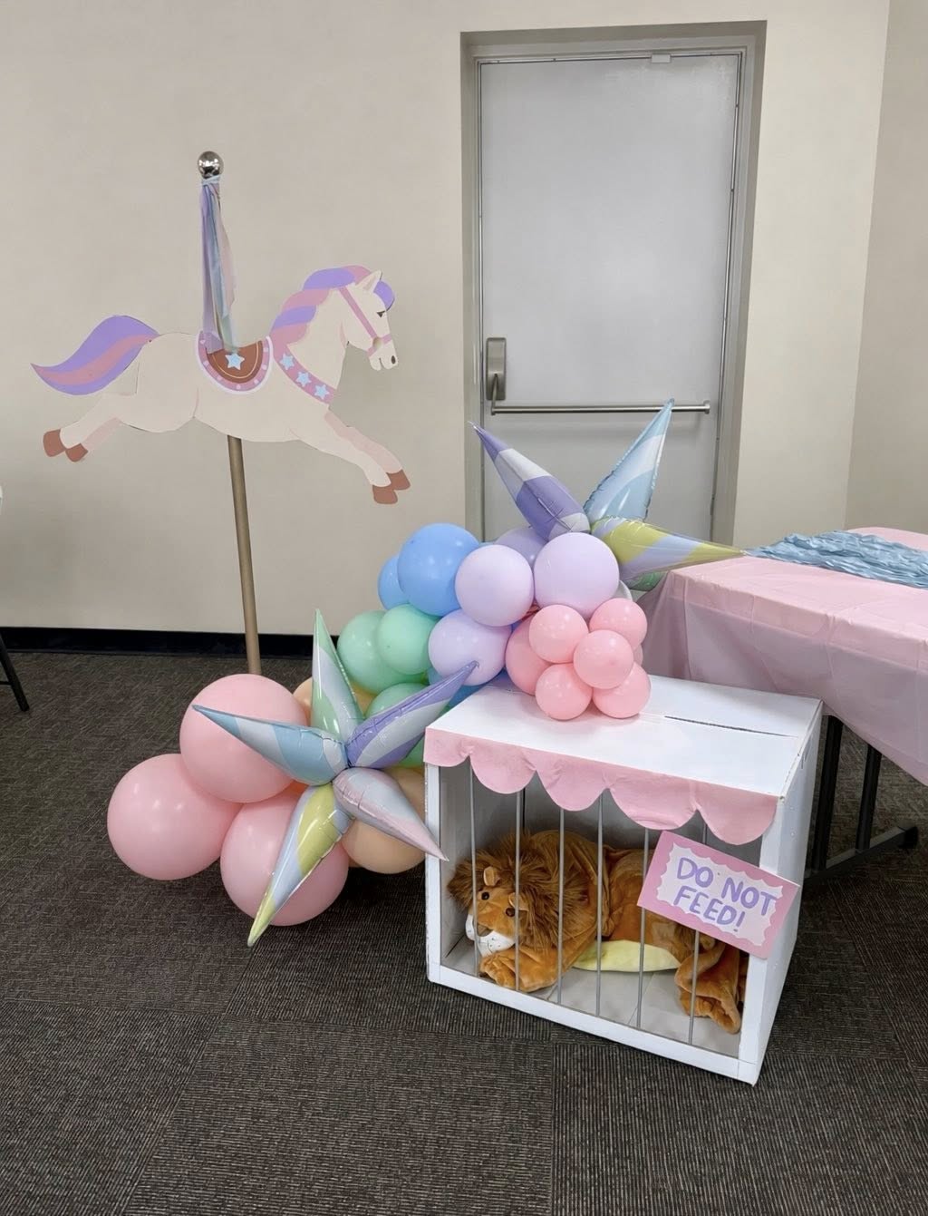 Circus party decor with pastel balloon arch, carousel horse cutout, and a toy lion in a cage with a Do Not Feed sign.