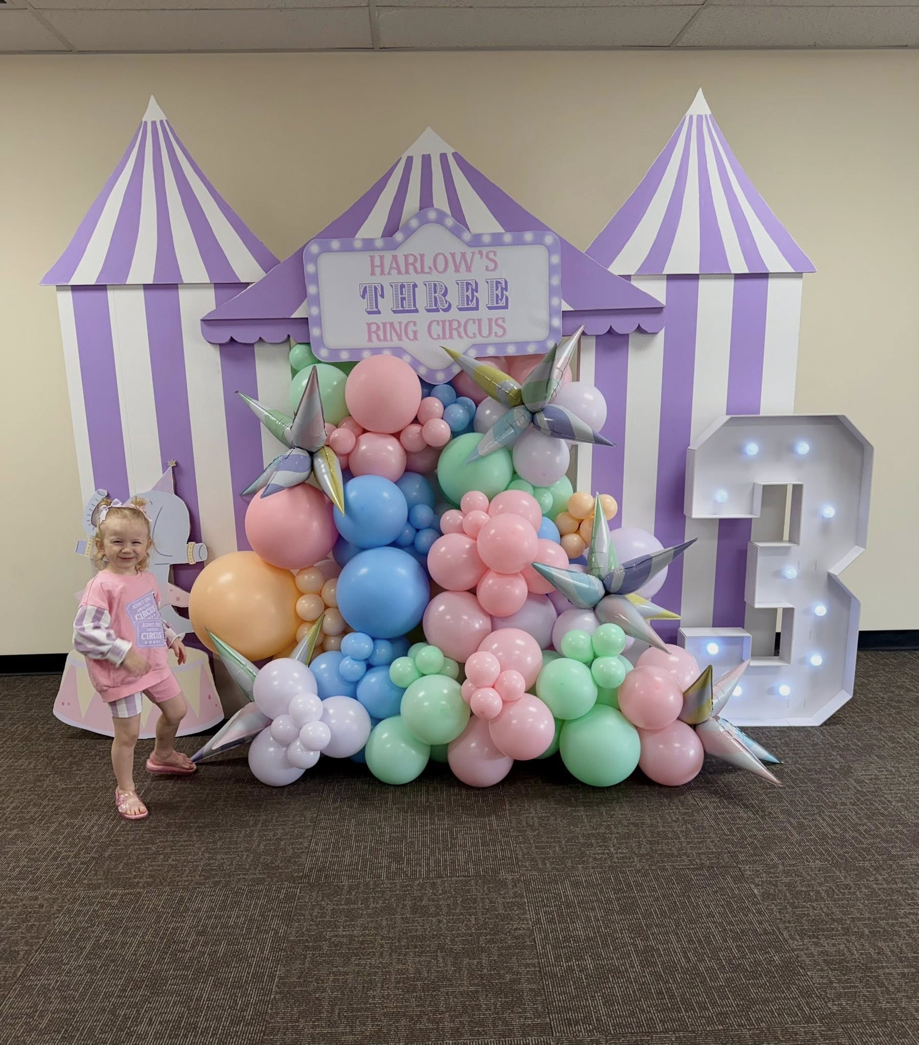 A young girl celebrating her 3rd birthday in front of a circus-themed backdrop with colorful balloon displays.