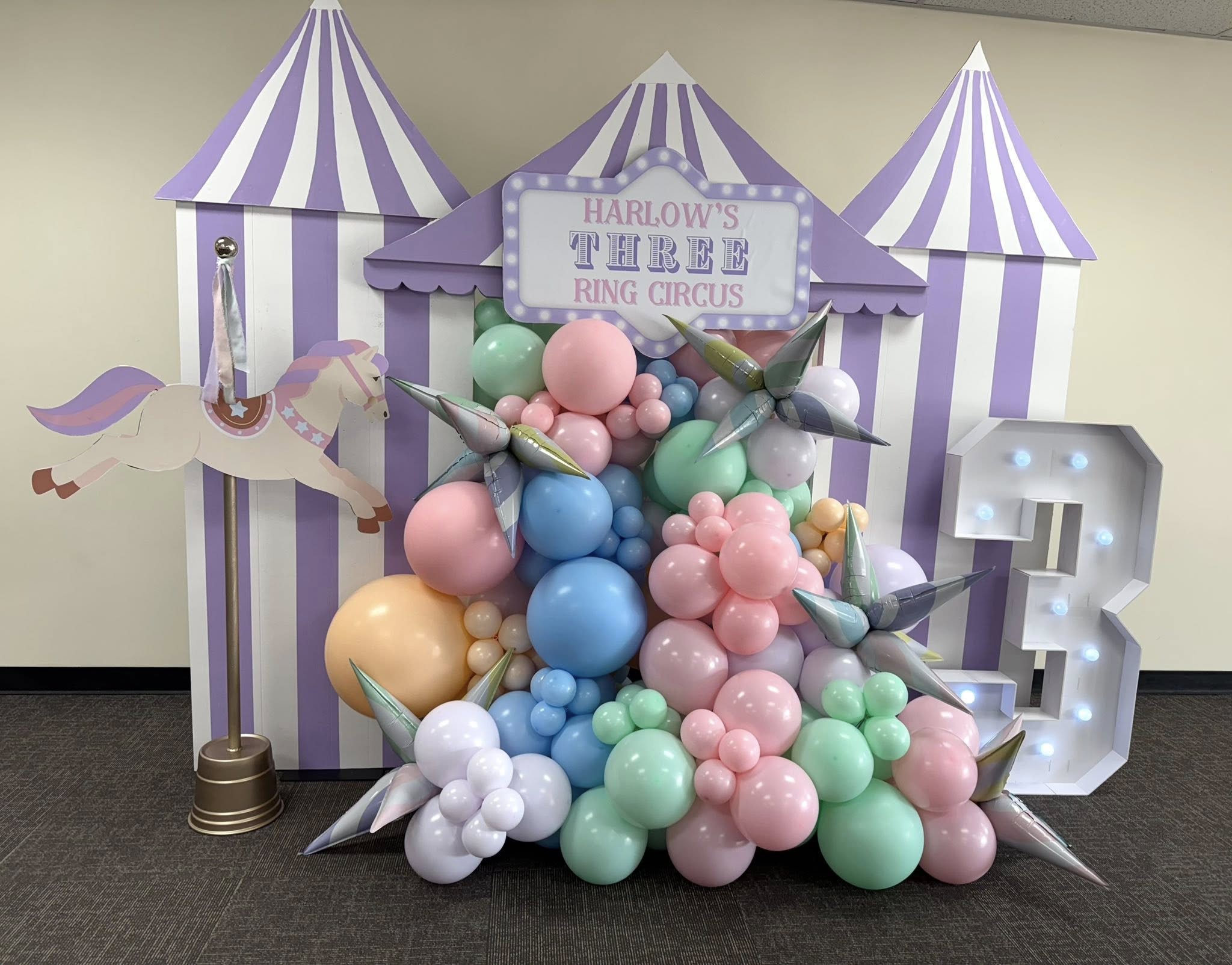 Circus-themed birthday party backdrop with pastel balloon garland, marquee sign for Harlow's Three Ring Circus, and a number 3 light.