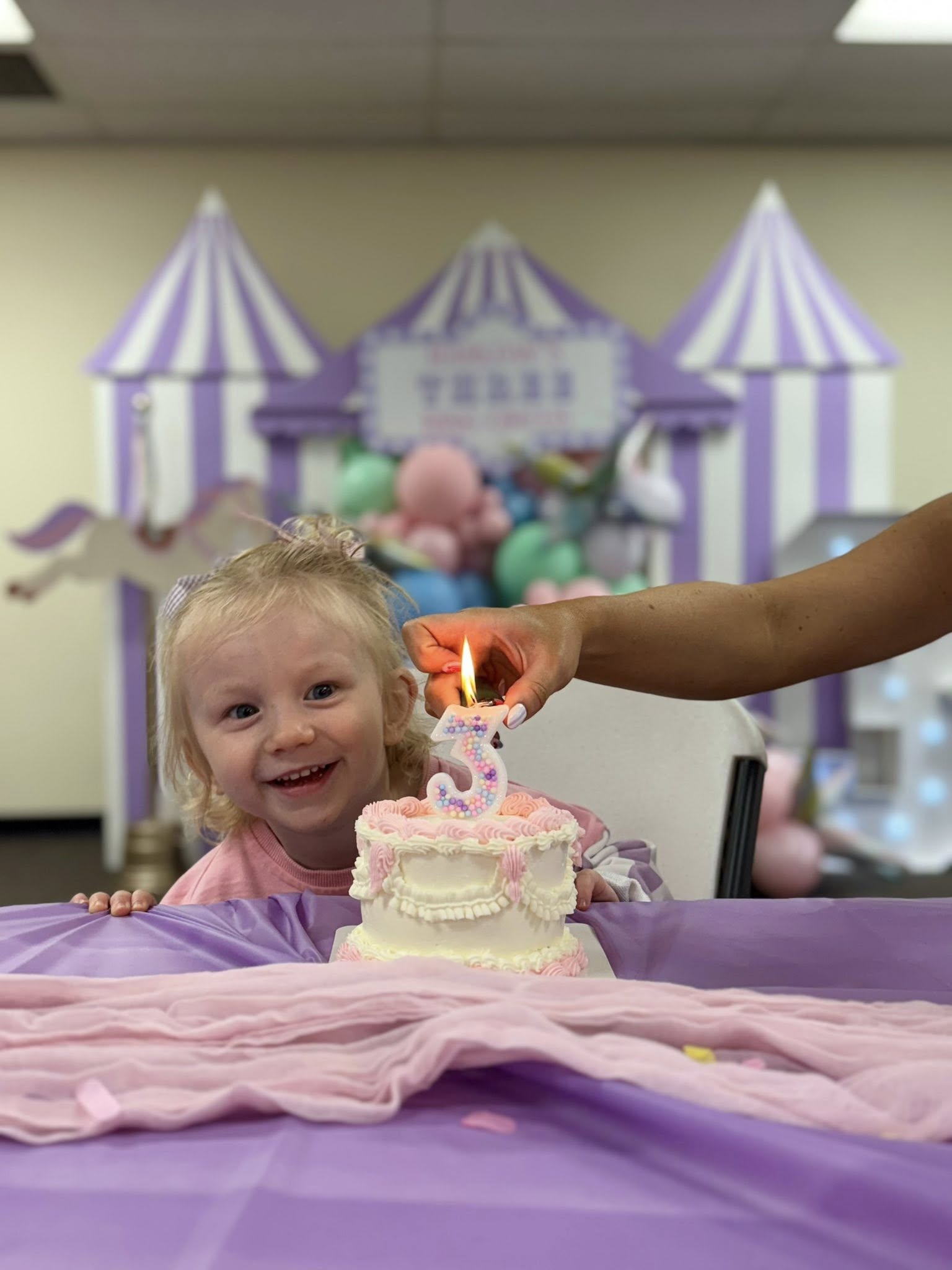 Toddler smiling behind a small birthday cake with a lit number three candle during a circus-themed party.