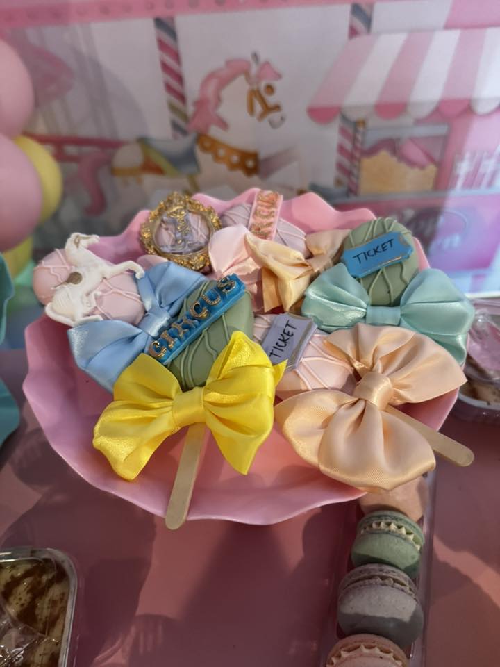 Set of circus-themed cake pops decorated with ribbons and ticket-shaped toppers on a pink serving tray.