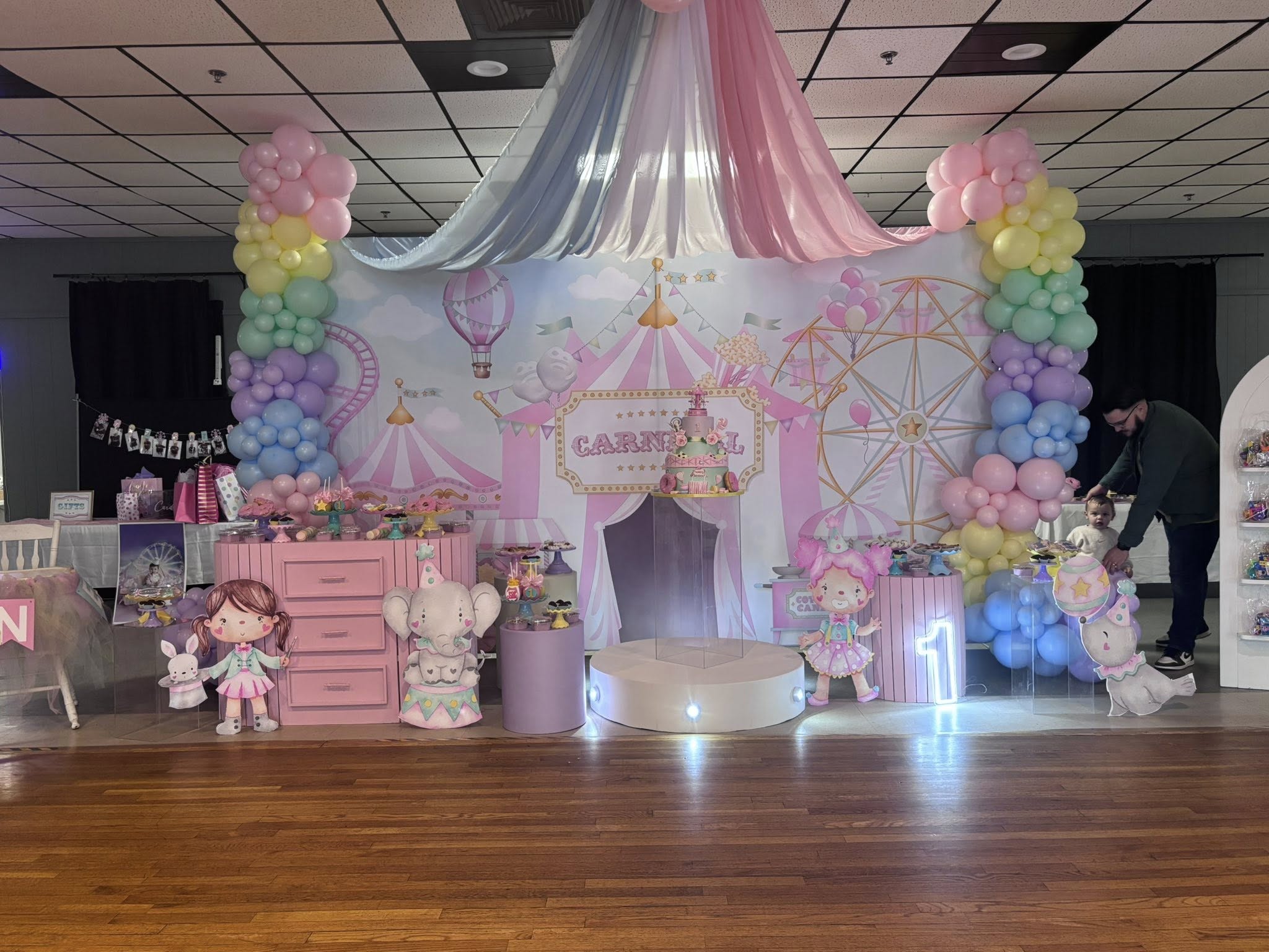 Carnival-themed first birthday party setup with pastel balloon pillars, carnival backdrop, and themed table decor.