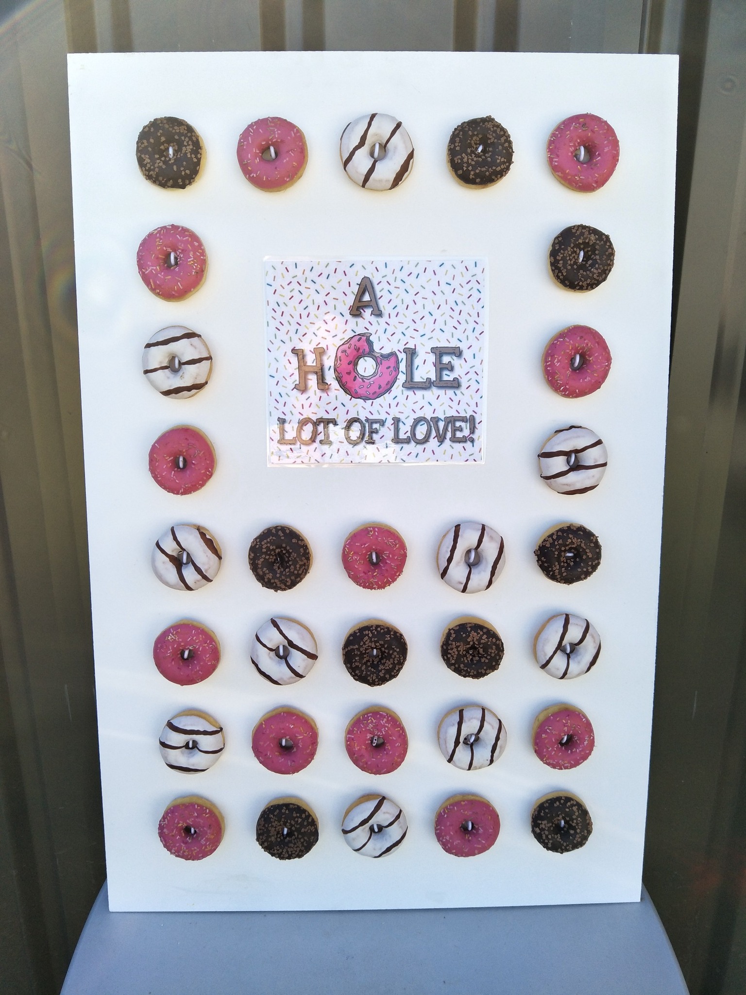 A white display board featuring rows of assorted pink and chocolate frosted donuts with a sign reading A Hole Lot Of Love.