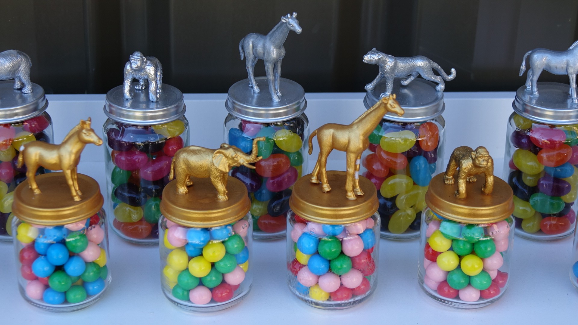 Glass candy jars with metallic animal figurines on lids, filled with colorful jelly beans and gumballs.