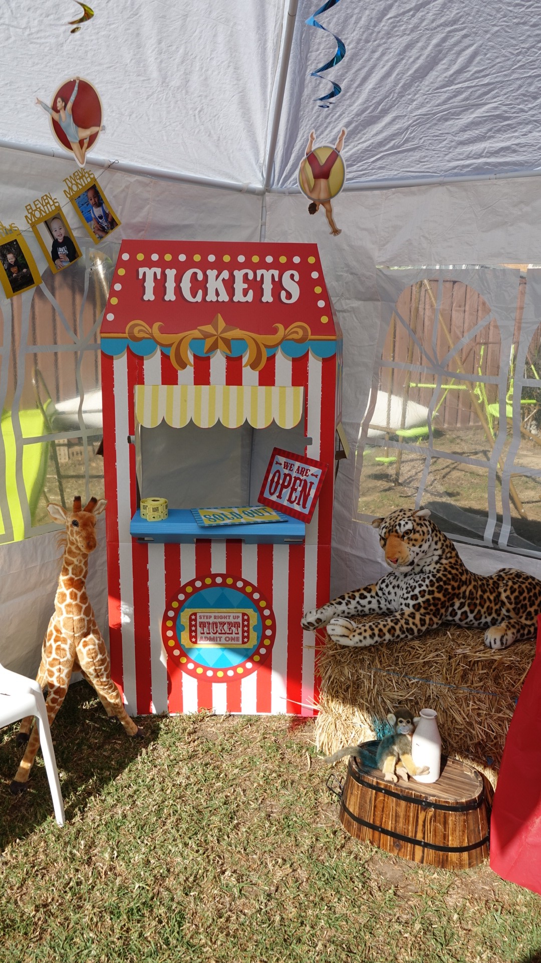 Cardboard circus ticket booth decoration flanked by stuffed giraffe and leopard toys on hay bale.