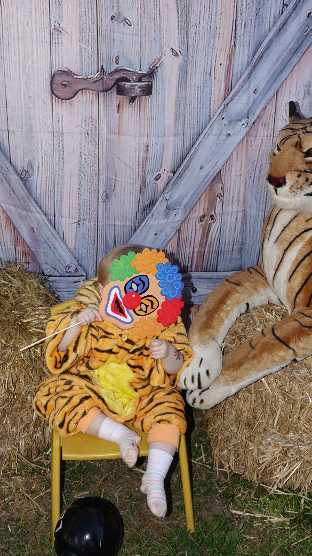 Toddler in a tiger costume holding a clown mask in front of their face, sitting on a chair next to a large plush tiger.