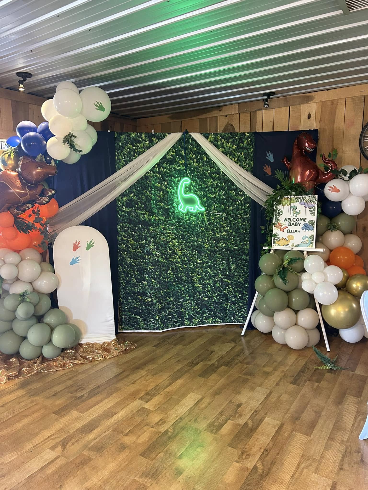 Dinosaur themed baby shower backdrop with greenery, balloons, and a Welcome Baby Elijah sign.