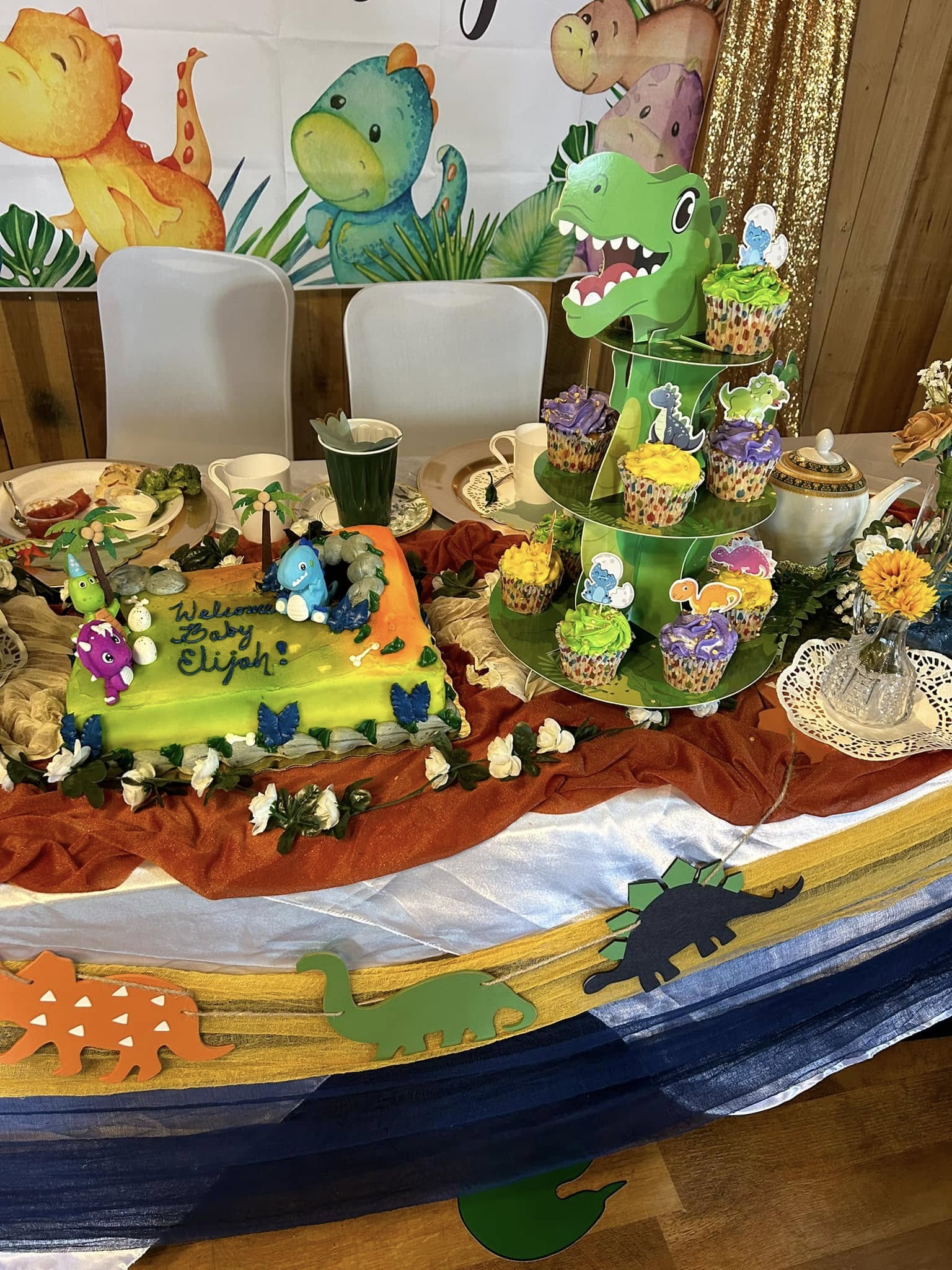 Dinosaur-themed baby shower decor featuring a 'Welcome Baby Elijah' cake and a tiered cupcake stand with toppers.
