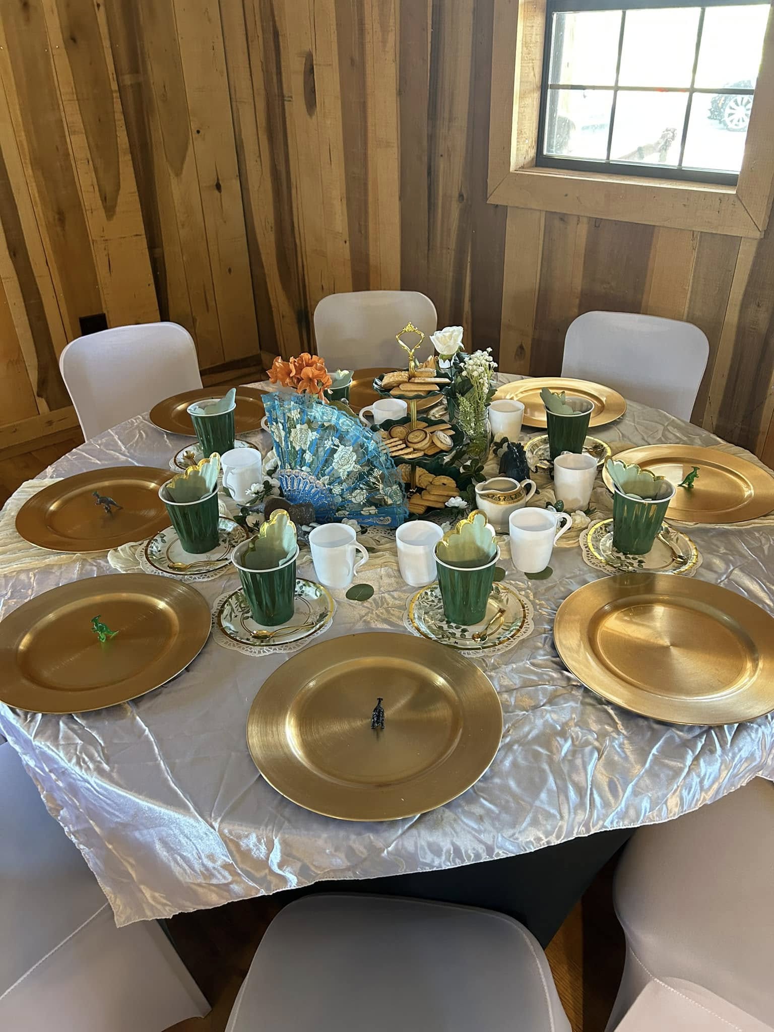 Table set for a tea party featuring gold chargers, green cups, and small toy dinosaurs as party favors.