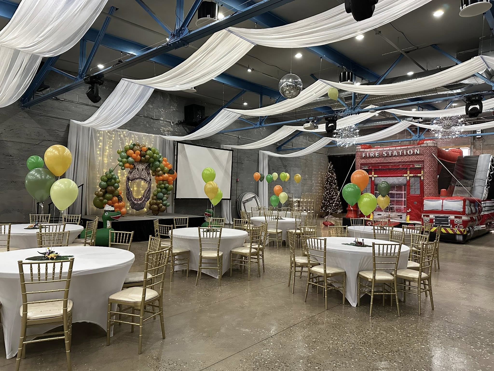 Event space decorated for a dinosaur and fire station theme, featuring round tables with balloons