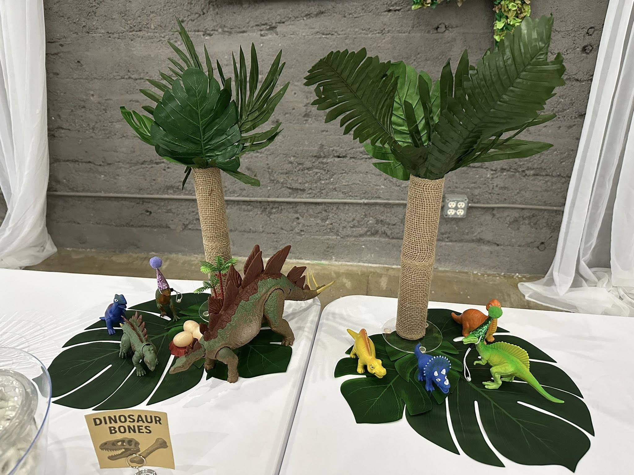 Dinosaur themed party table centerpiece with toy dinosaurs, palm tree decorations, and a dinosaur bones sign.