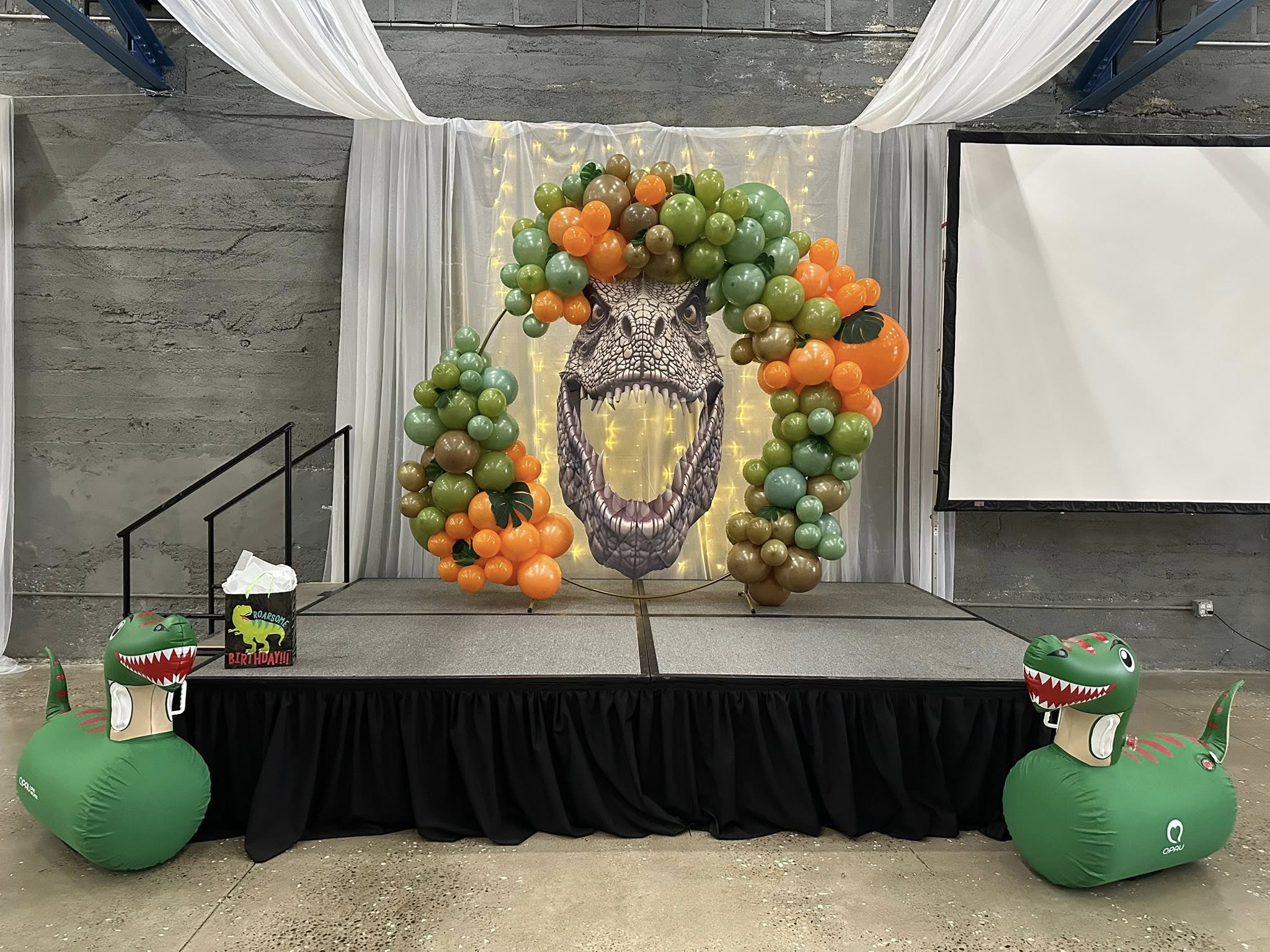 Dinosaur themed party stage with balloon arch surrounding an open-mouthed T-Rex cutout and inflatable dinosaurs