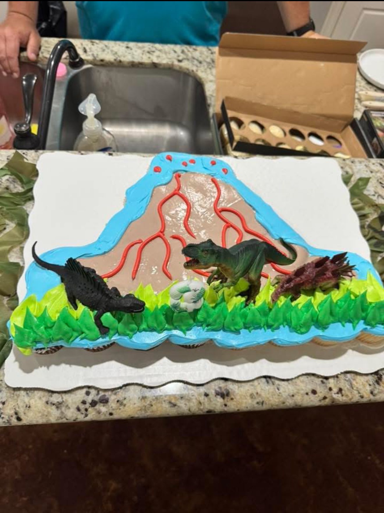 Cupcake cake decorated as a volcano with icing lava and three plastic dinosaur figurines on top.