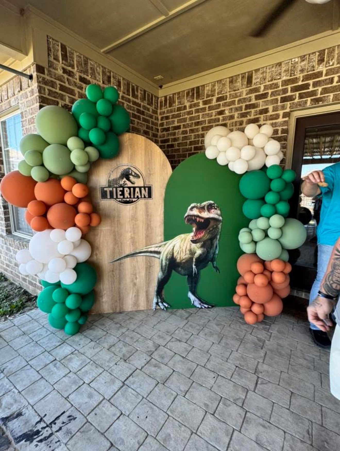 Dinosaur themed birthday party backdrop featuring T-Rex cutout and green, orange, and white balloon arch.
