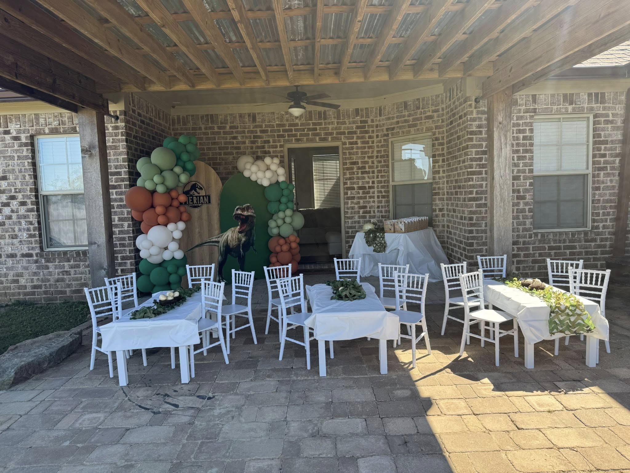 Dinosaur themed birthday party outdoor setup with white chairs, dining tables, and green and orange balloon arch