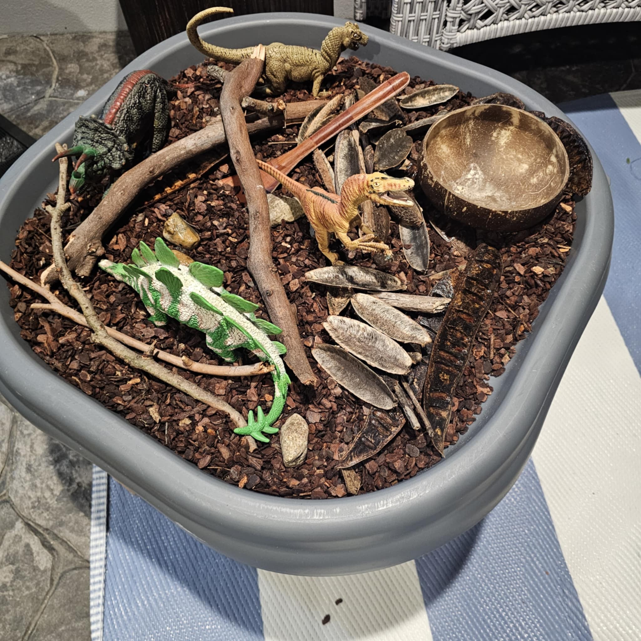 Dinosaur sensory bin filled with wood chips, twigs, dried seed pods, and small toy dinosaurs.