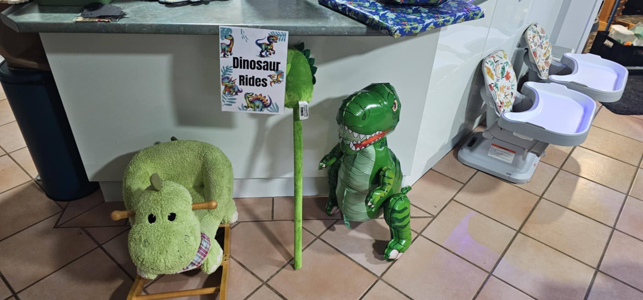 Dinosaur themed play area with a green plush rocker, a toy dinosaur stick, and a T-Rex balloon under a sign.