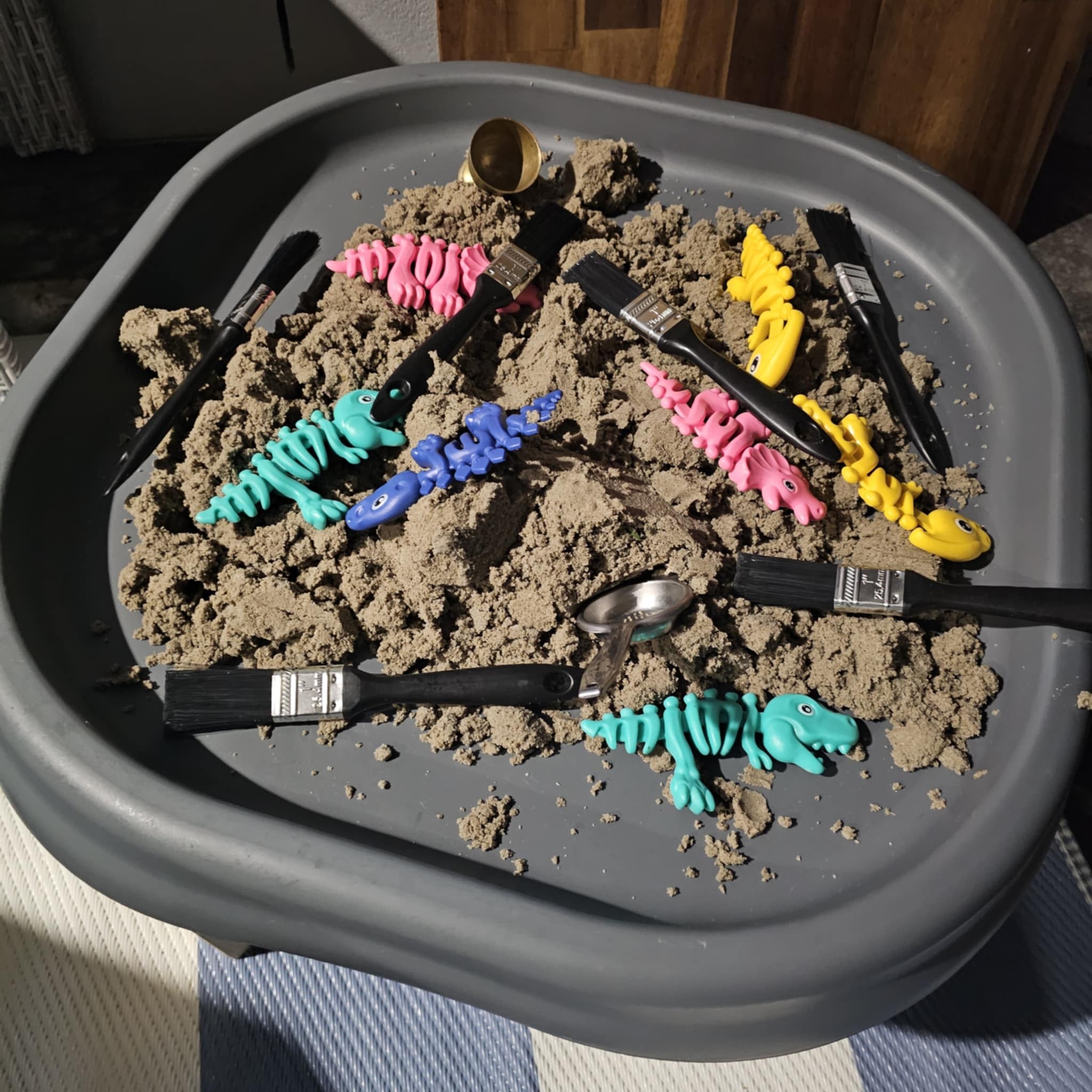 Sensory tray filled with sand, toy dinosaur skeletons, and paintbrushes for a dino excavation activity.