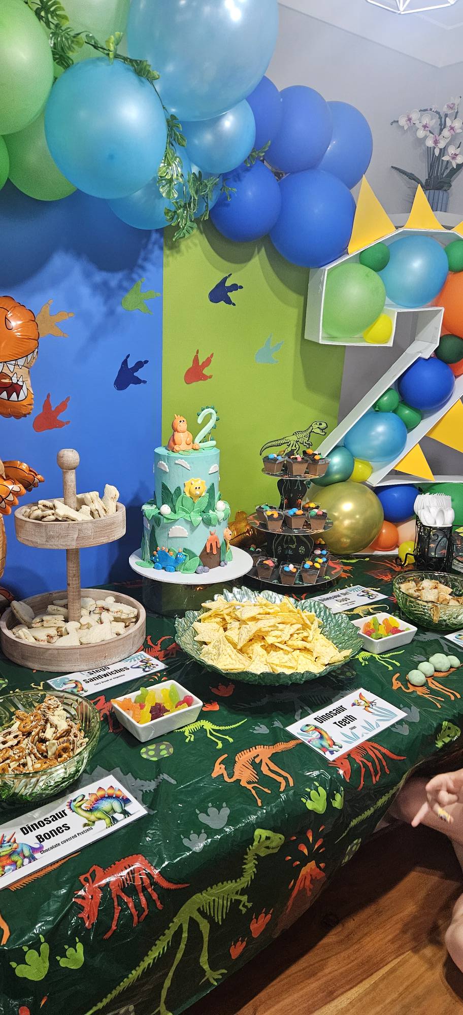Dinosaur themed birthday party table with a two-tier cake, snacks, and balloon decorations.