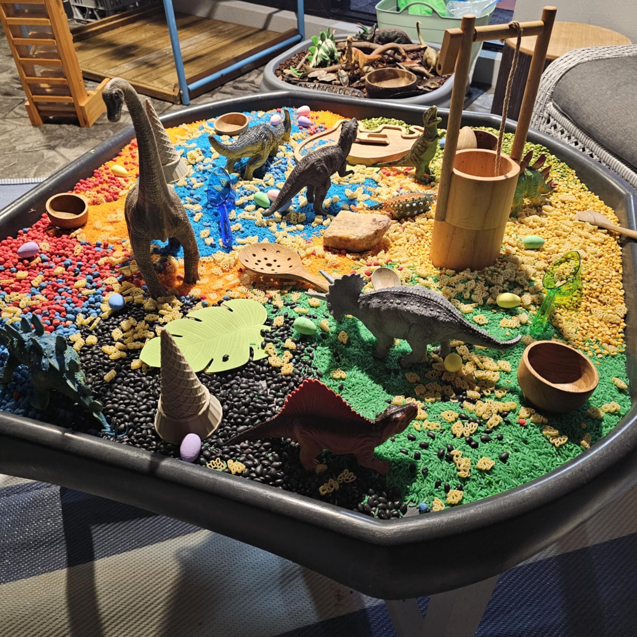Dinosaur themed sensory play tray with colorful dyed rice, black beans, plastic dinosaur figures, and wooden accessories.