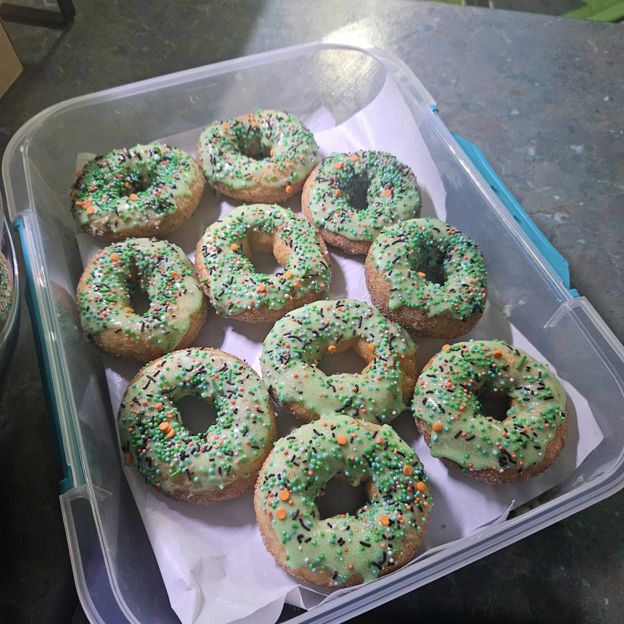 Homemade donuts with light green icing and colorful sprinkles arranged in a plastic container