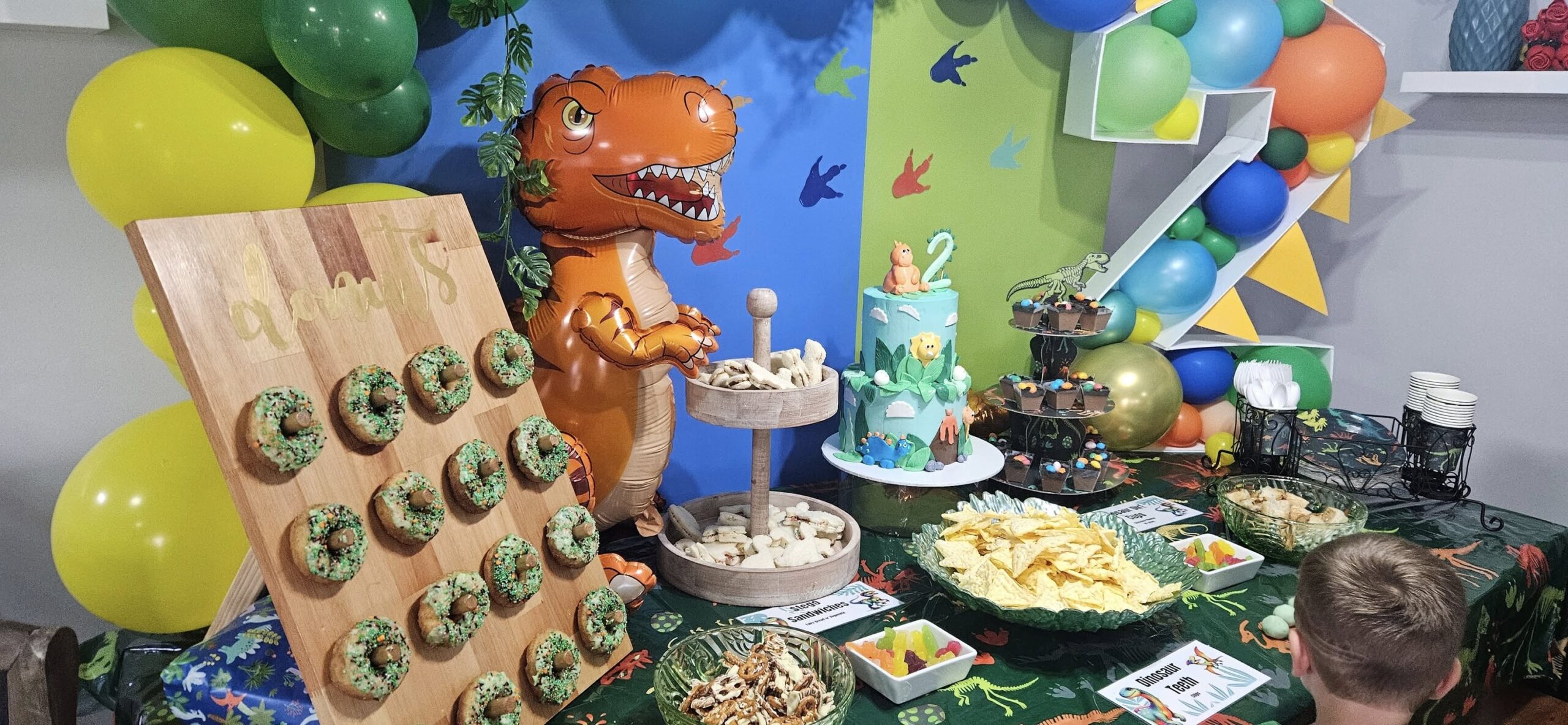 Dinosaur themed birthday dessert table with a t-rex balloon, cake, and donut wall.
