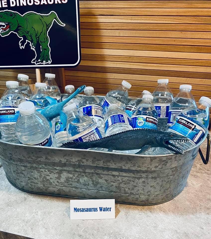 Galvanized metal tub filled with Kirkland bottled water, decorated with toy dinosaurs and a Mosasaurus Water label.