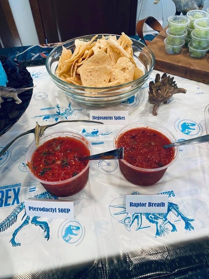 Dinosaur themed party snacks including tortilla chips labeled Stegosaurus Spikes and salsa labeled Raptor Breath.
