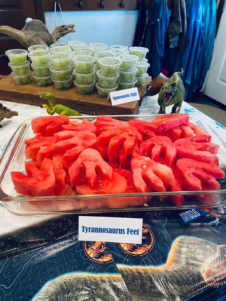 Dinosaur birthday party food display featuring watermelon cut into dinosaur footprints labeled as Tyrannosaurus Feet.