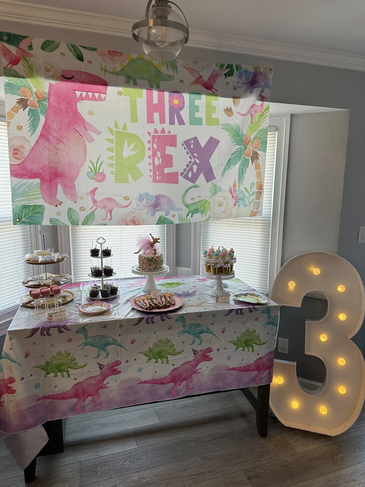 Dinosaur-themed third birthday party table setup with a Three Rex banner, decorated cake, and lighted number 3.