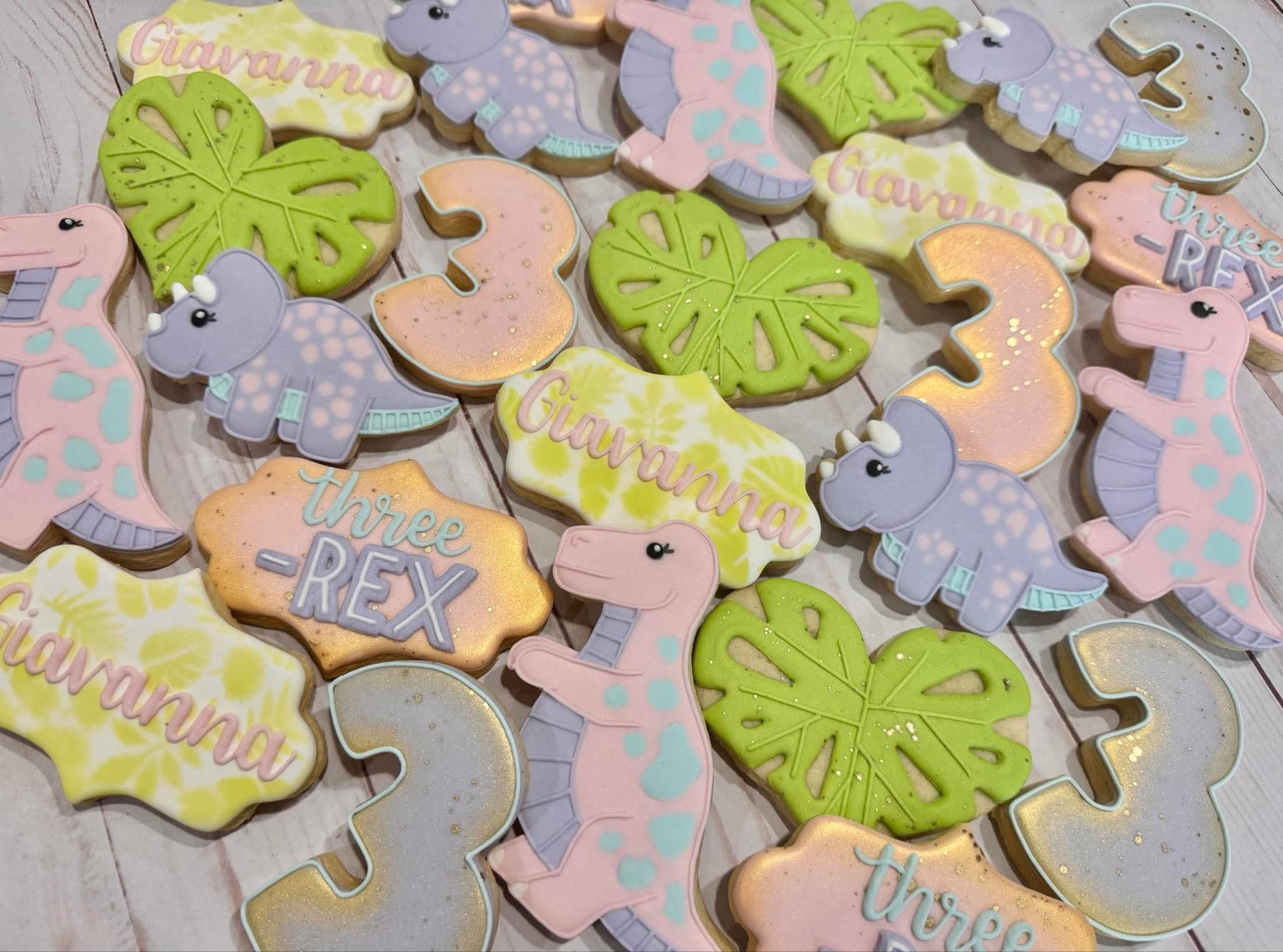 Custom dinosaur-themed cookies shaped like number 3s, triceratops, t-rexes, palm leaves, and name plaques.