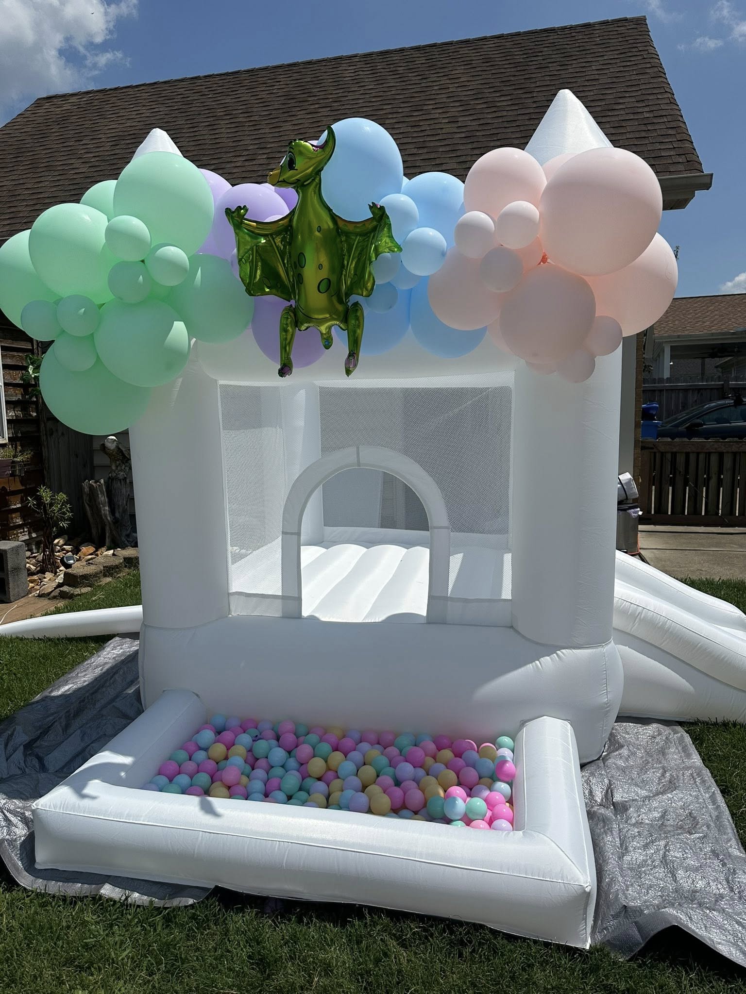 White bounce house with a ball pit in front, decorated with pastel balloons and a green dinosaur balloon