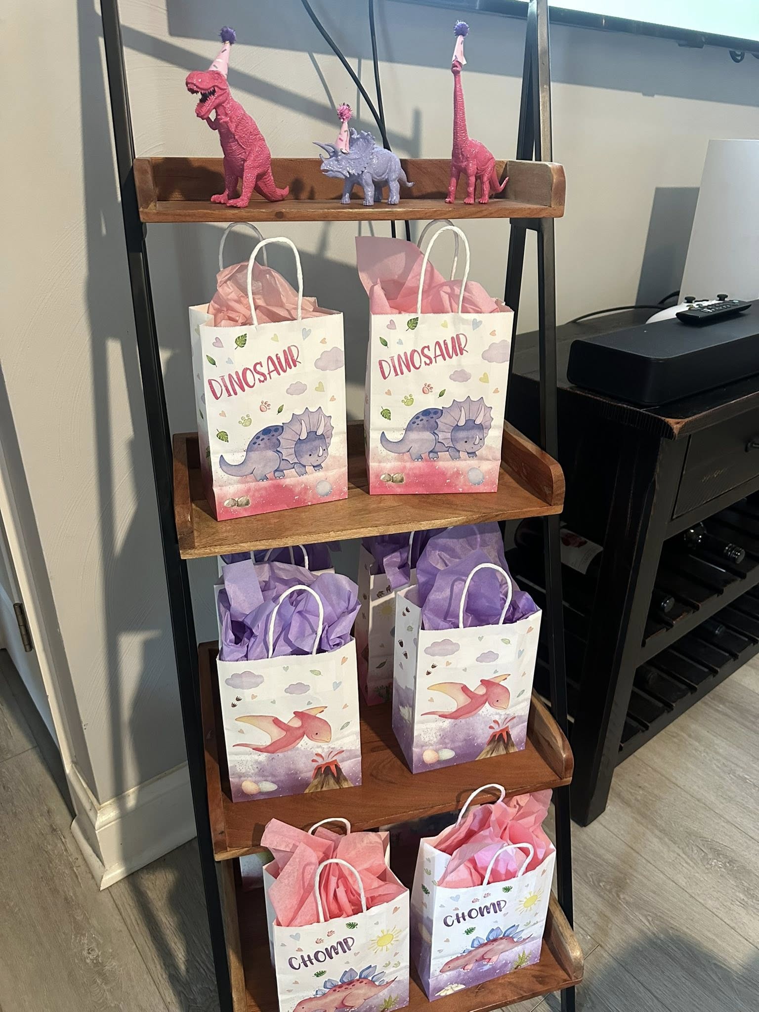 Dinosaur theme party favor bags with pink and purple tissue paper arranged on a wooden tiered shelf