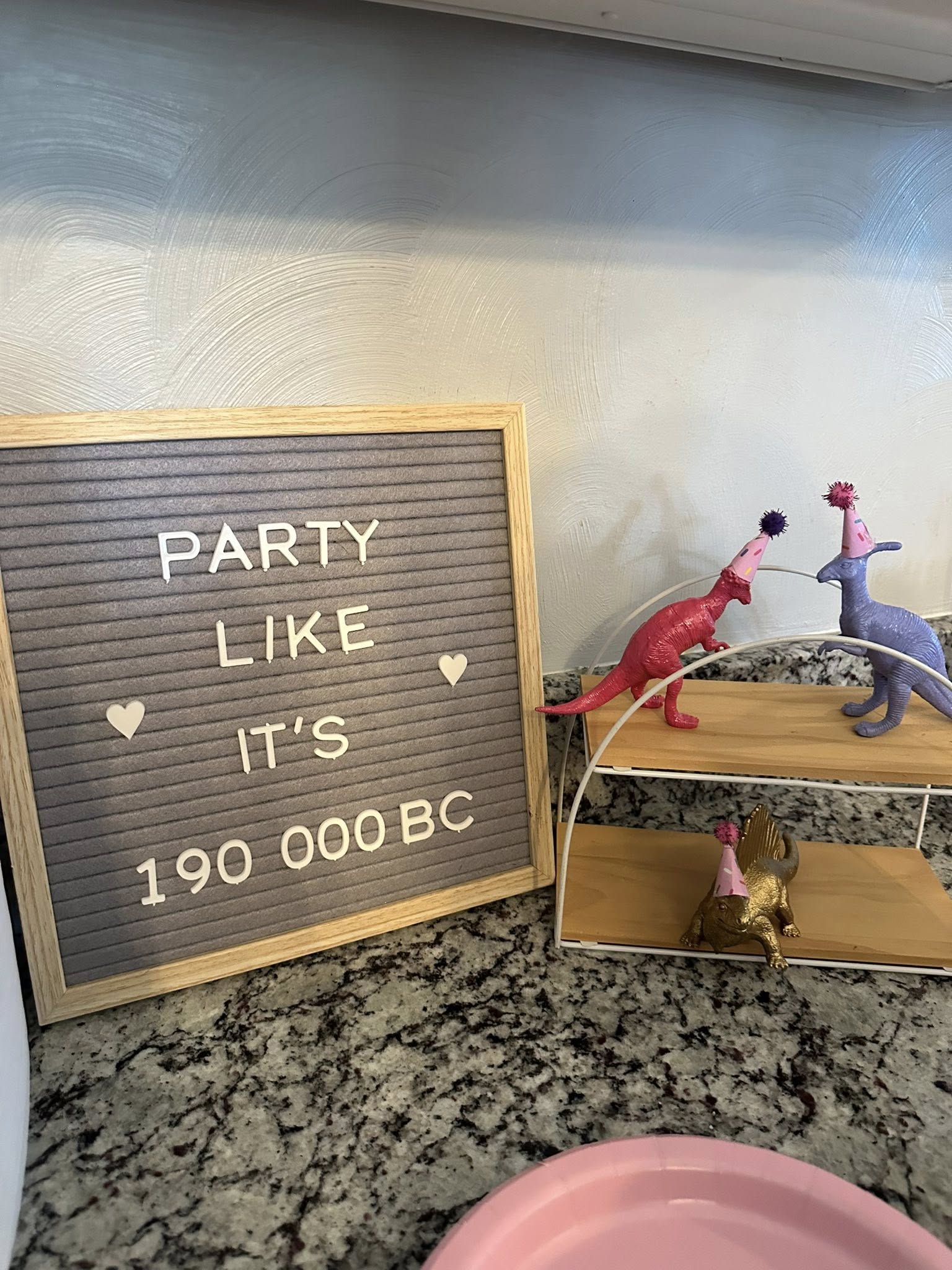 Felt letter board reading Party Like It's 190,000 BC next to toy dinosaurs wearing party hats on a tiered stand.