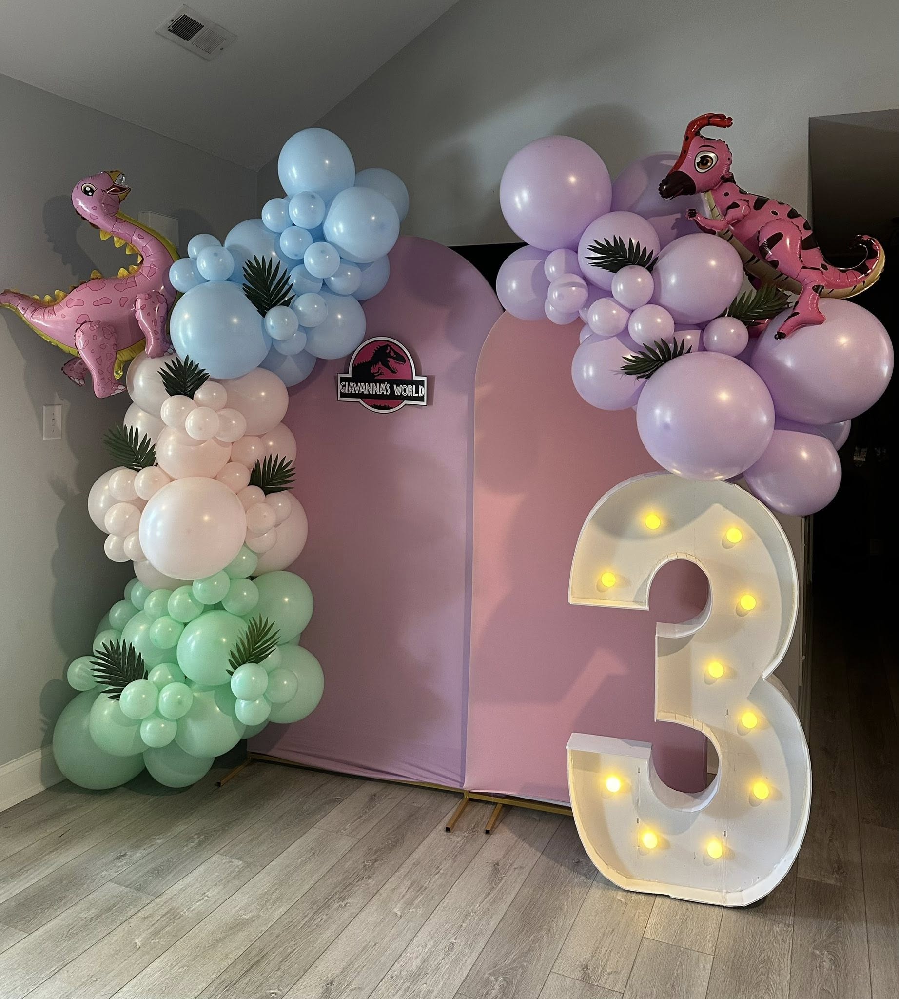 Dinosaur-themed third birthday party backdrop with pastel balloon garland and illuminated number 3