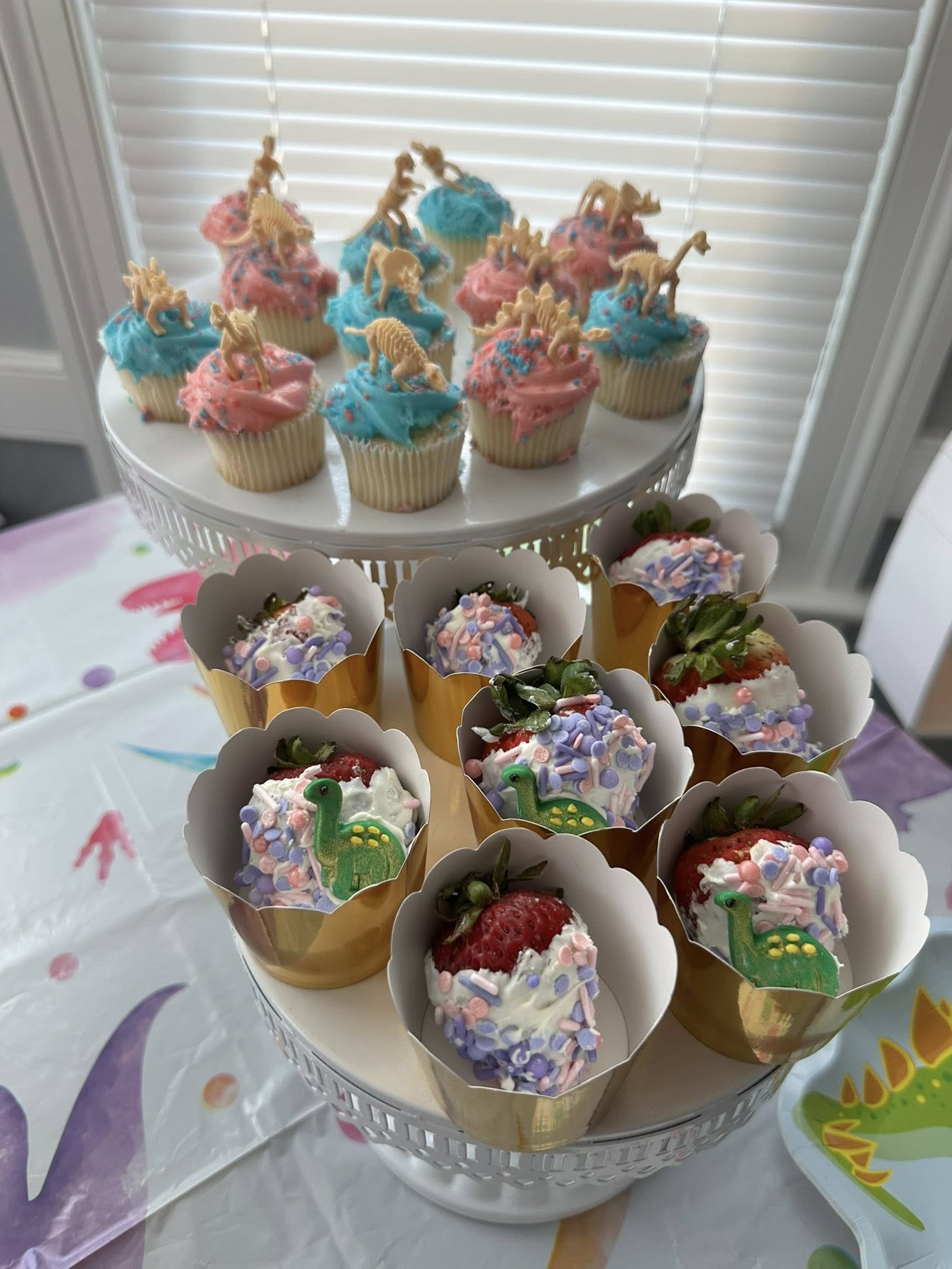 Tiered stand featuring dinosaur-topped cupcakes and chocolate-dipped strawberries with pastel sprinkles.
