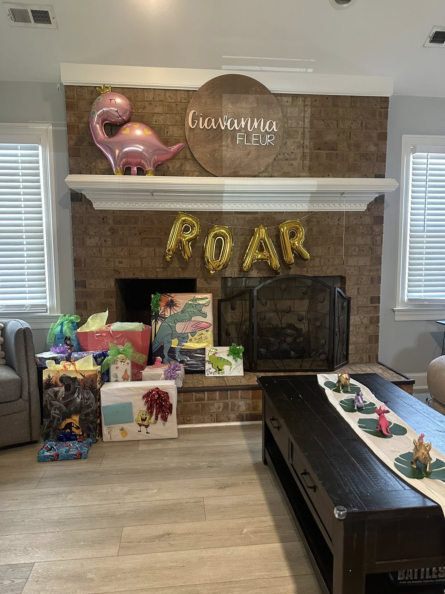 Dinosaur-themed birthday party decorations with a ROAR balloon banner over a fireplace featuring gifts and name sign