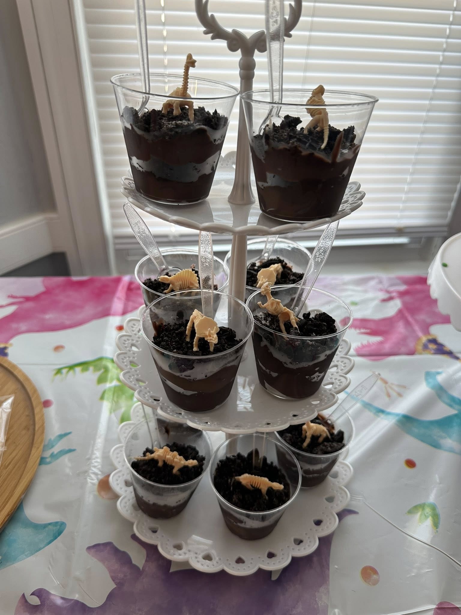 Tiered stand holding individual dessert cups topped with crushed chocolate cookies and plastic dinosaur toys.