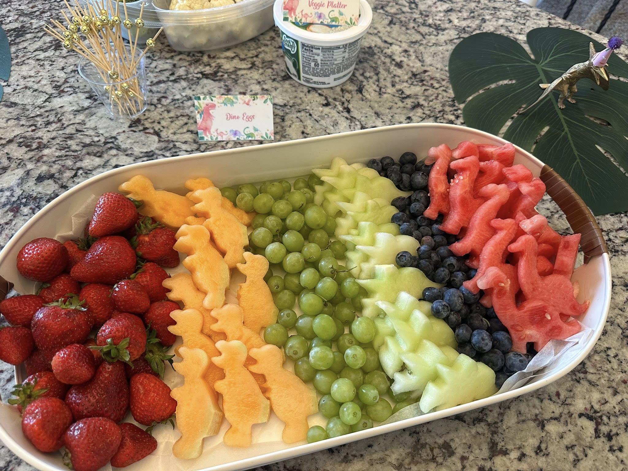 Dinosaur-themed fruit platter featuring melon shaped like dinosaurs, strawberries, grapes, and blueberries.