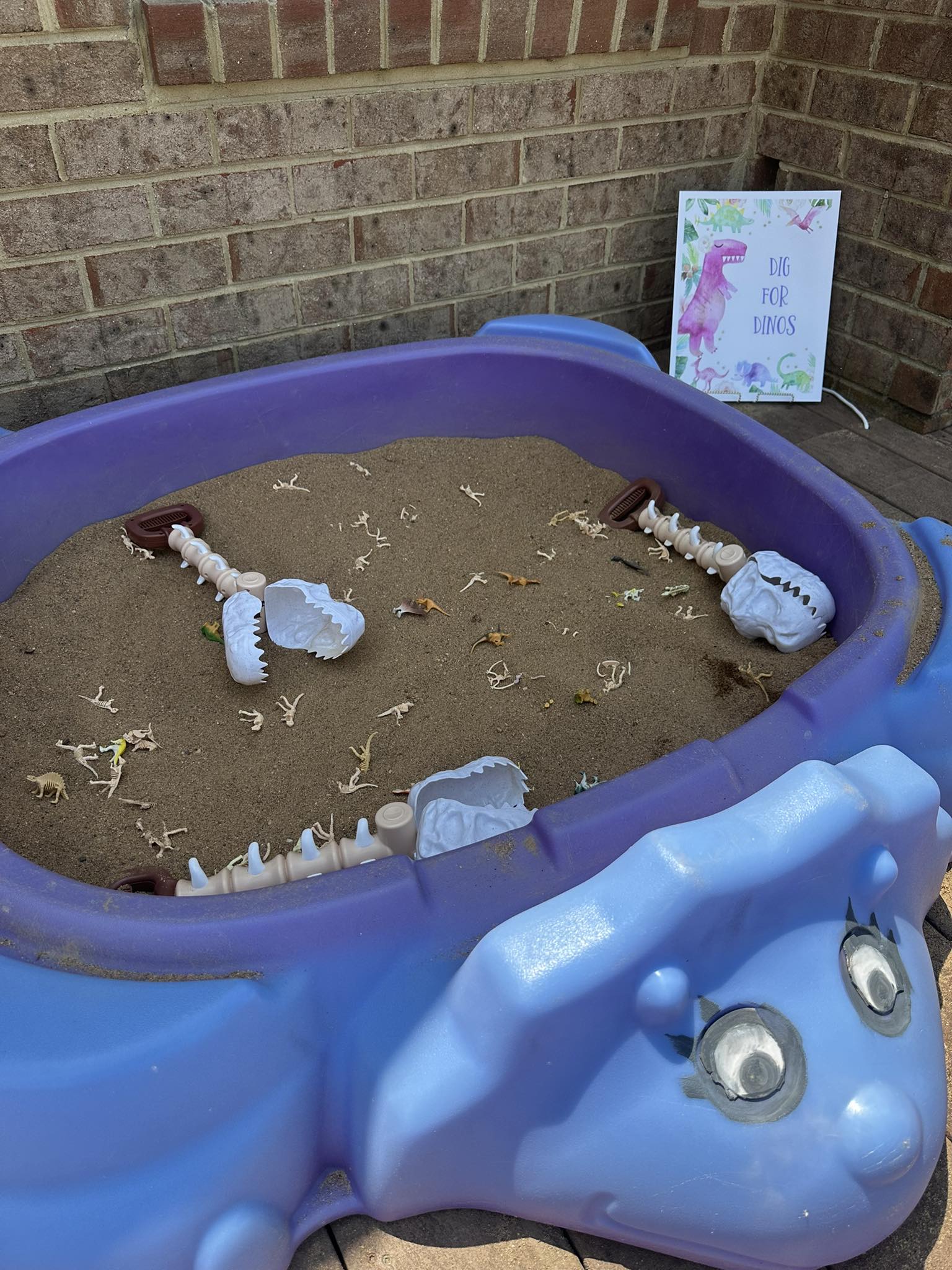 Blue dinosaur-shaped sandbox filled with sand, toy dinosaur skeletons, and a Dig for Dinos sign.