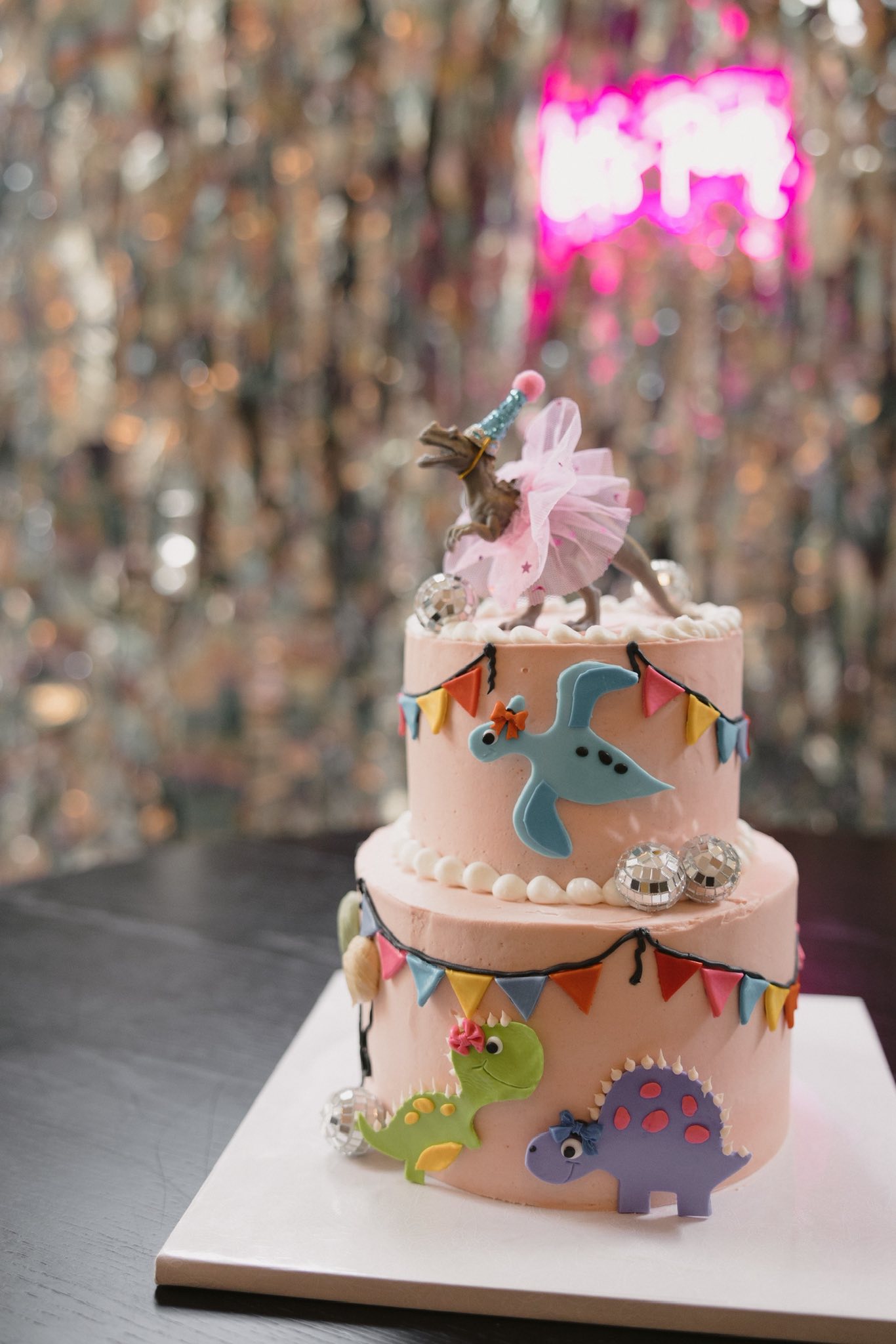 Two-tiered pink birthday cake topped with a T-Rex wearing a tutu, decorated with colorful dinosaur cutouts and mini disco balls.