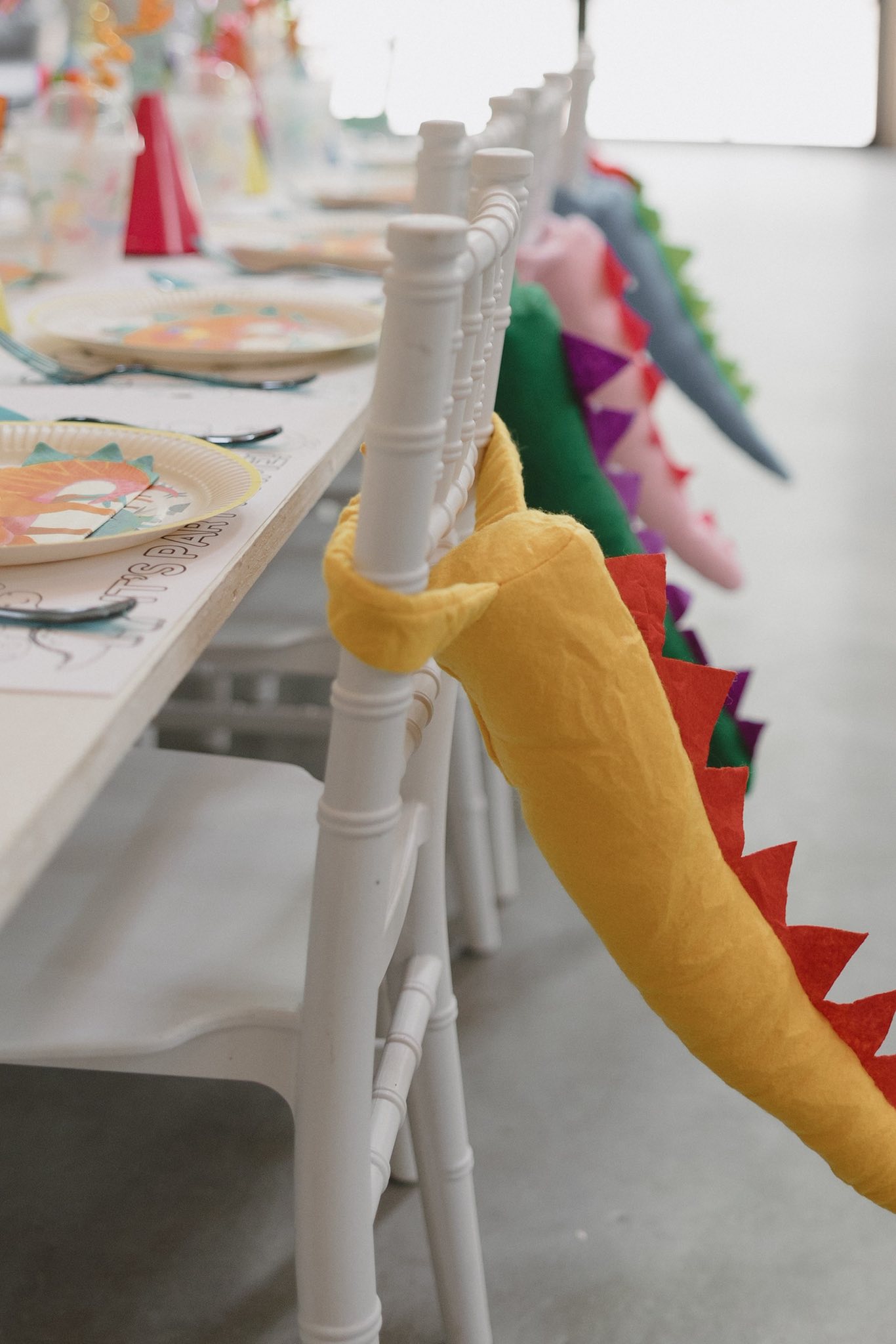 White chairs decorated with colorful plush dinosaur tails for a themed party table setting.