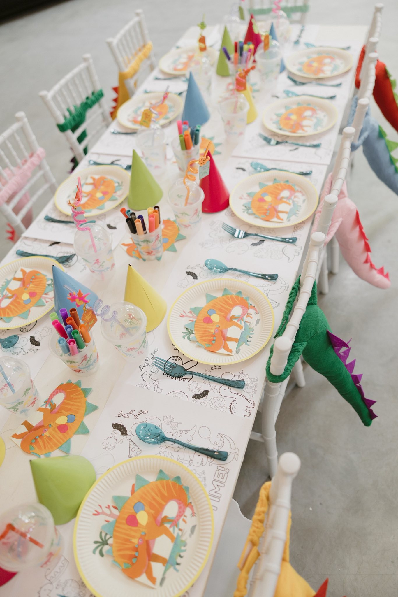 Dinosaur-themed party table set with plates, party hats, cups with markers, and dinosaur tail chair backs.