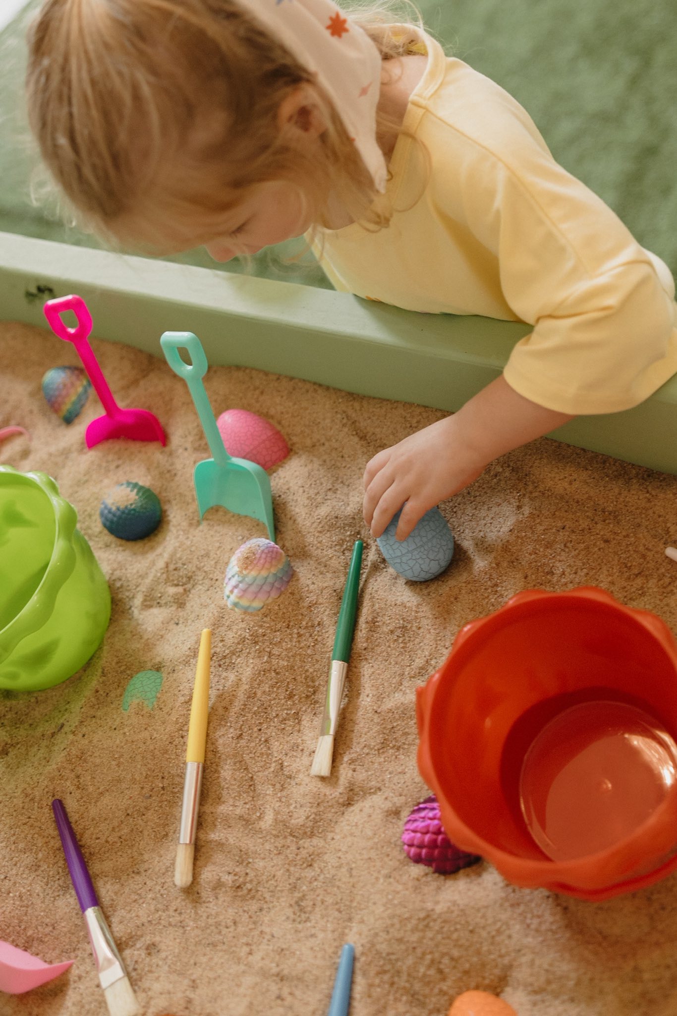Toddler playing in a sandbox with colorful mini shovels, paintbrushes, and textured egg-shaped toys.