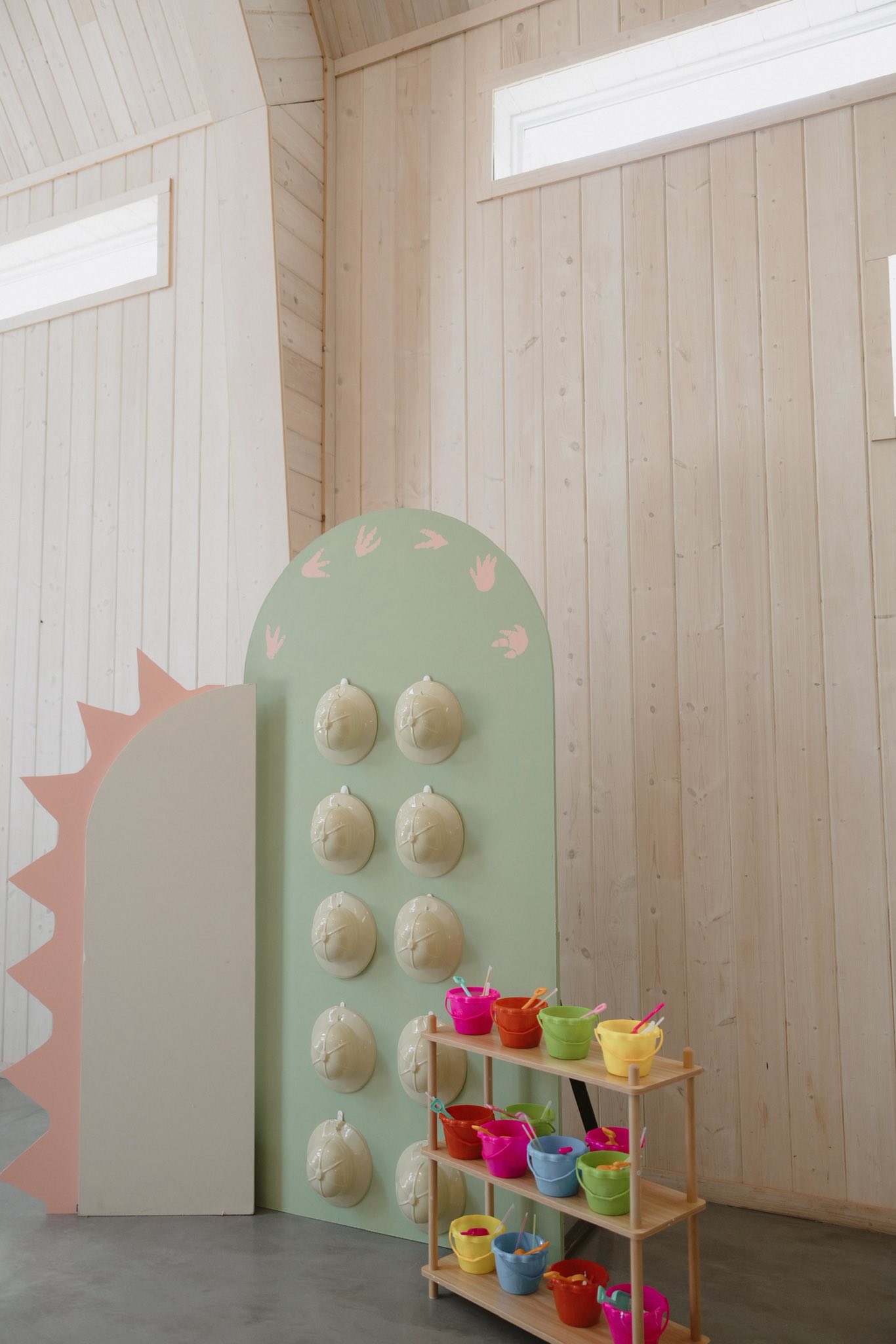 Dinosaur-themed display board with hanging explorer hats and a wooden shelf filled with colorful play buckets.