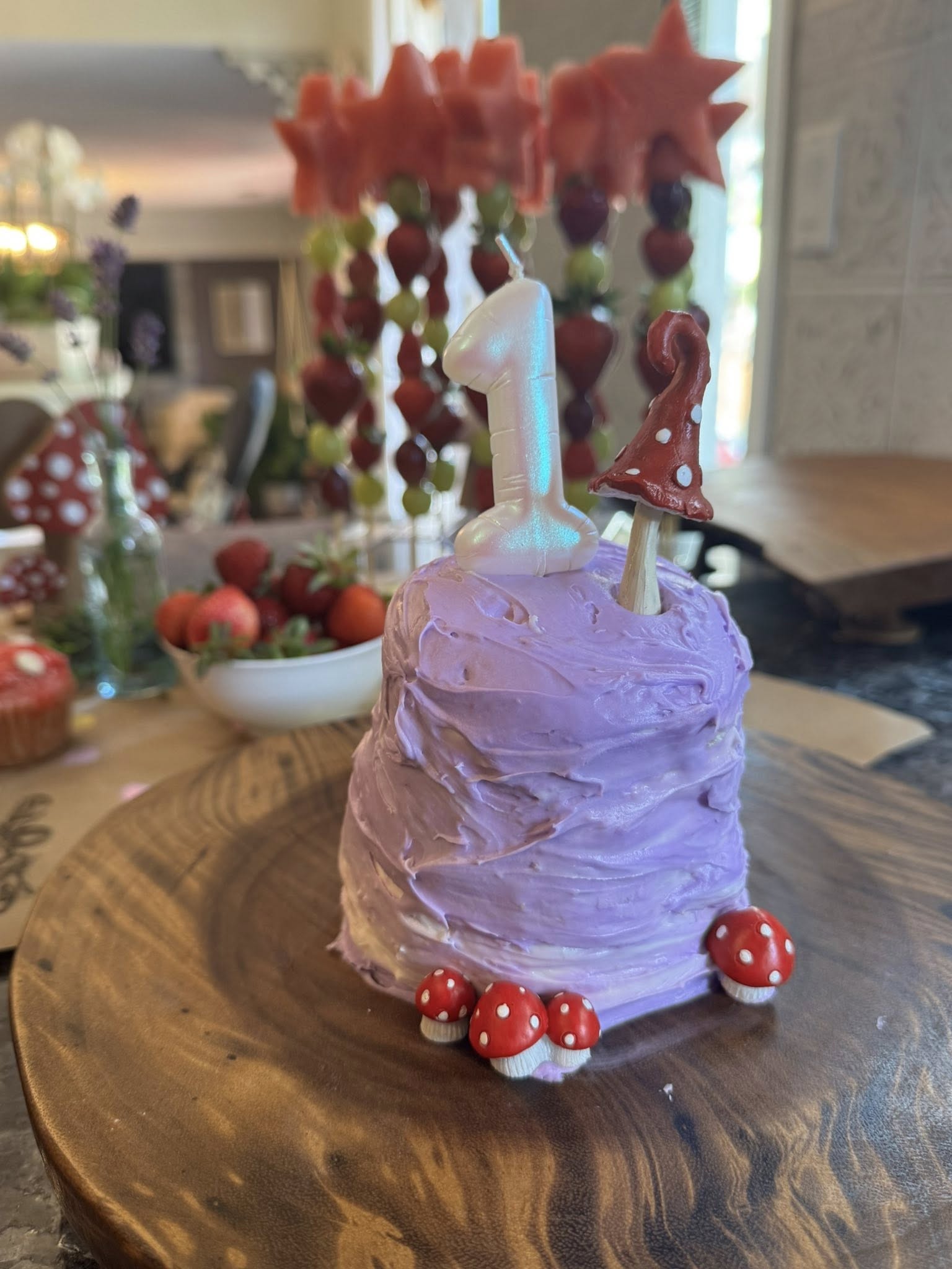 Small round cake with lavender frosting, topped with a number one candle and toadstool mushroom decorations.