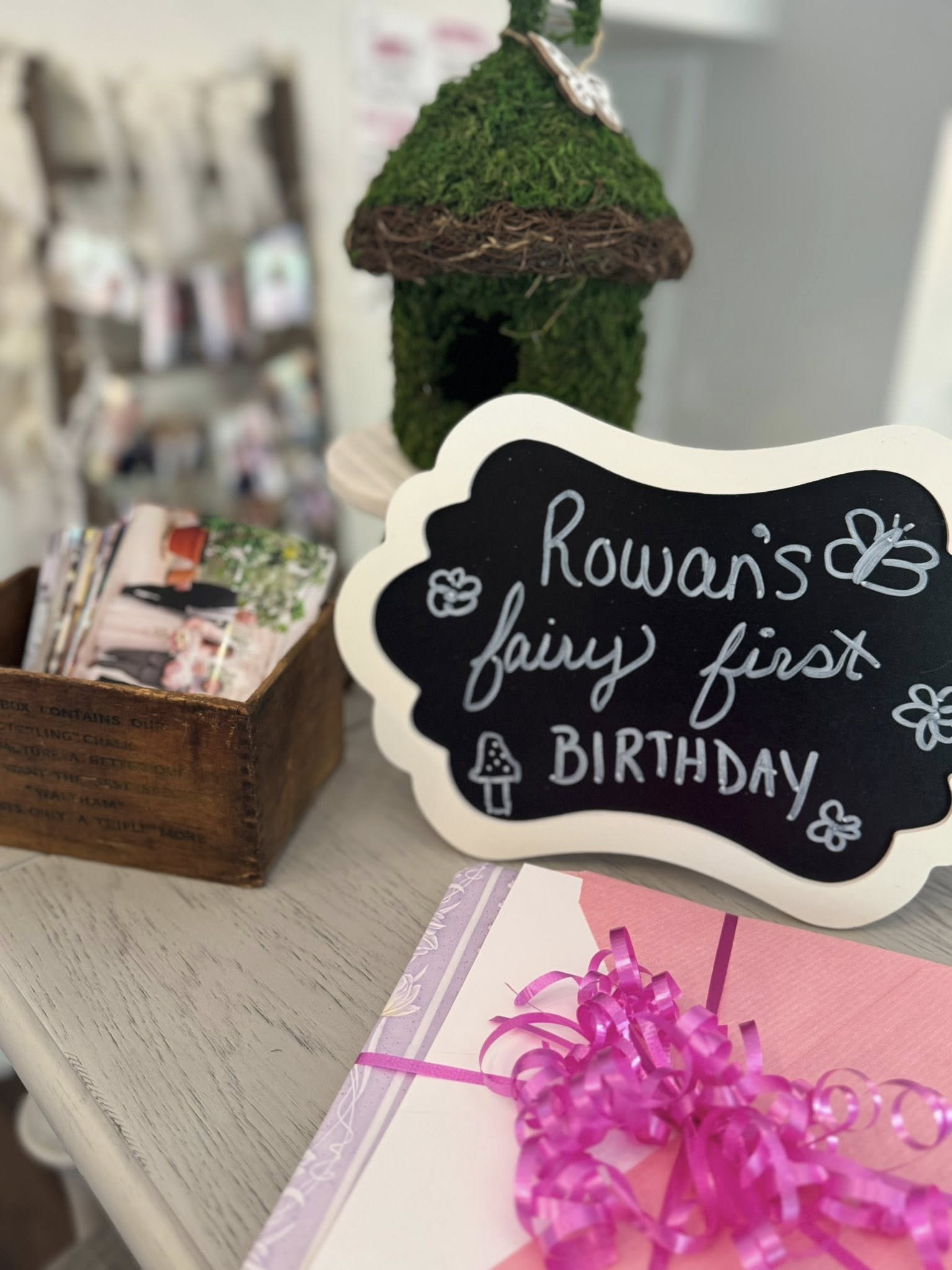 Blackboard table sign for Rowan’s fairy-themed first birthday party with a moss-covered birdhouse and party favors.