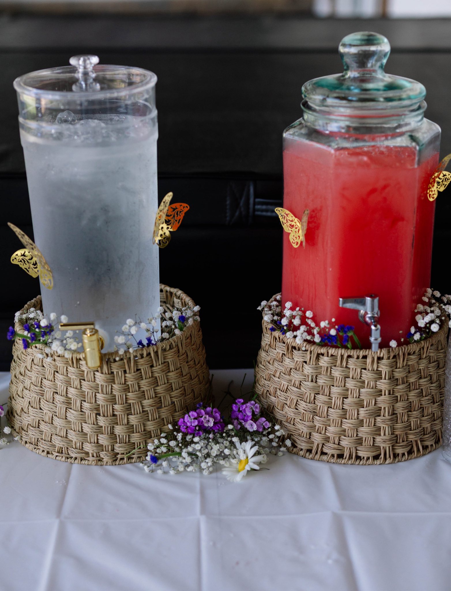 Two glass beverage dispensers decorated with butterfly accents and sitting in woven baskets with small flowers.