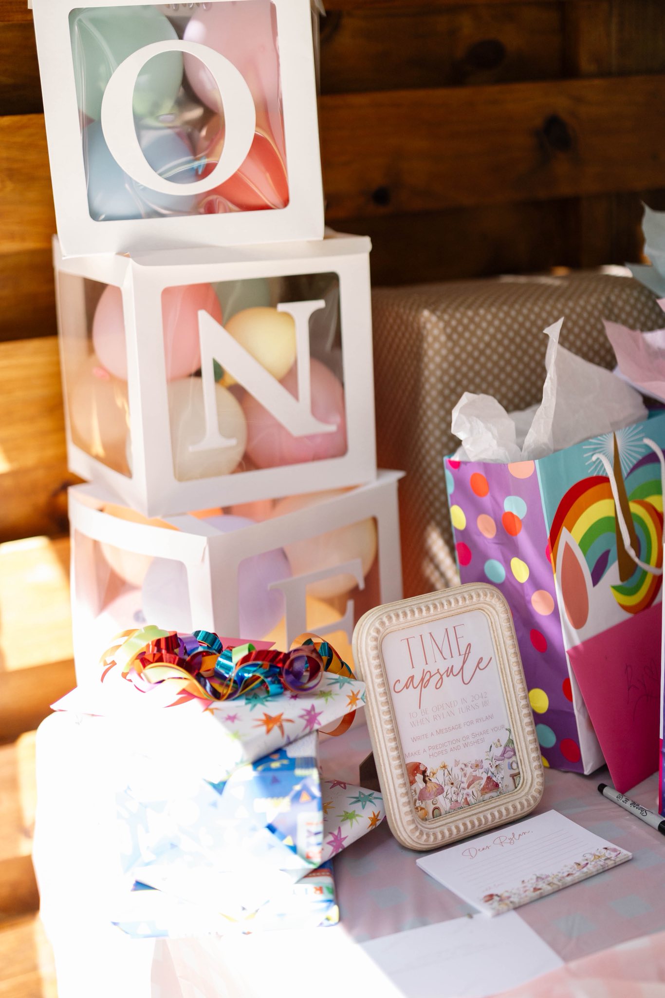 A stack of boxes spelling ONE filled with pastel balloons next to a Time Capsule sign and birthday gifts.