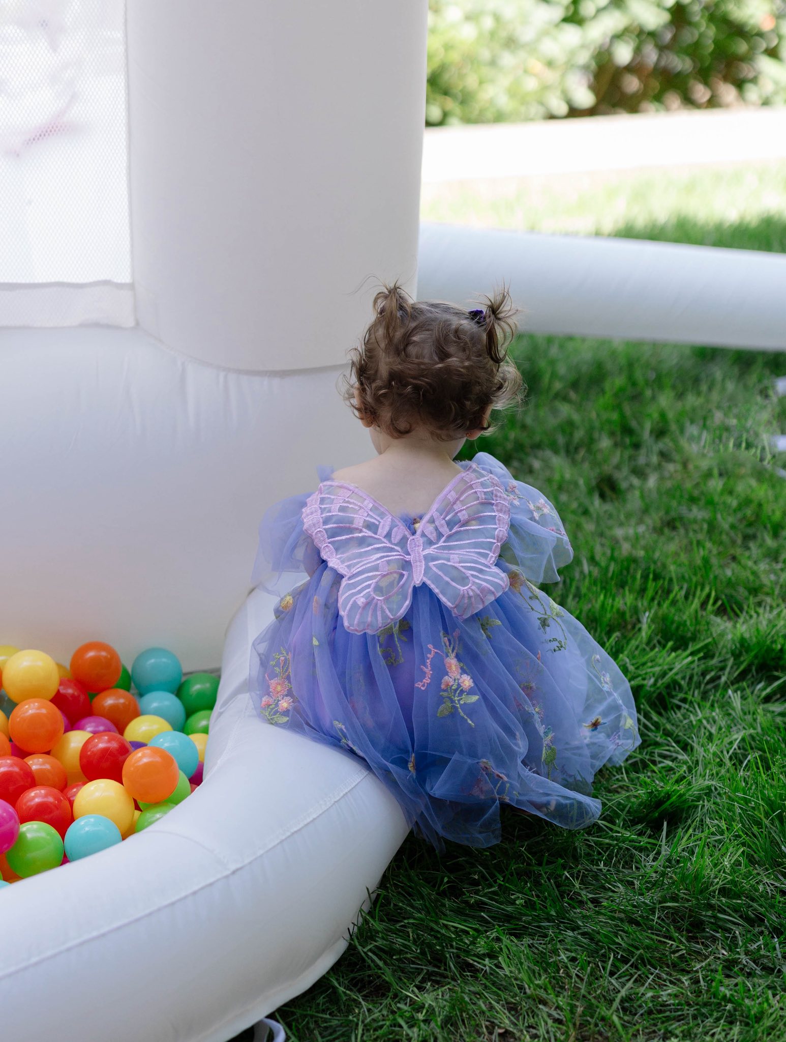 Toddler wearing a blue floral fairy dress with butterfly wings sitting next to a white ball pit outside.